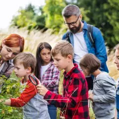 Grupa dzieci i dorosłych ogląda rośliny w lesie, jedno dziecko używa lupy.