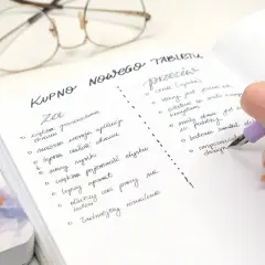 Ręka pisze notatkę w zeszycie: "Kupno nowego tabletu". Widoczne okulary i fragment innego notatnika.