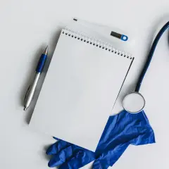 Lekarz przygotowuje dokumentację medyczną, notując informacje dotyczące usprawiedliwiania nieobecności w szkole rozporządzenie. Na stole leżą stetoskop, termometr, notatnik i rękawiczki.