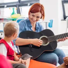Nauczycielka gra na gitarze dla dzieci, tworząc zindywidualizowaną ścieżkę kształcenia. Przykłady takiej nauki to muzyka.