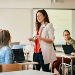 Uśmiechnięta kobieta, w trakcie przygotowania do zawodu nauczyciela, prowadzi lekcję dla grupy studentek przy laptopach.