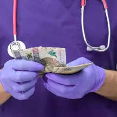 Pielęgniarka w fioletowym uniformie i rękawiczkach liczy pieniądze. Ile zarabia pielęgniarka po studiach?