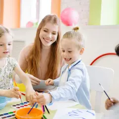 Nauczycielka z uśmiechem pomaga dzieciom malować farbami w przedszkolu.
