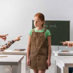 Smutna dziewczynka w brązowej sukience stoi sama, podczas gdy inne dzieci wskazują na nią palcami. To obrazek, który mógłby posłużyć jako opinia o uczniu do sądu, pokazując jego izolację.