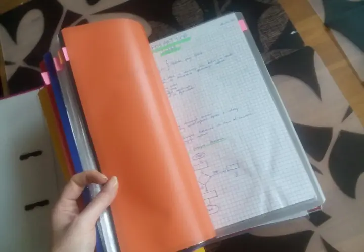 Ręka trzyma pomarańczową okładkę segregatora, odsłaniając zeszyt z notatkami i schematem. Widać nagłówek "FORMATYKA" i zasady prowadzenia zeszytu.