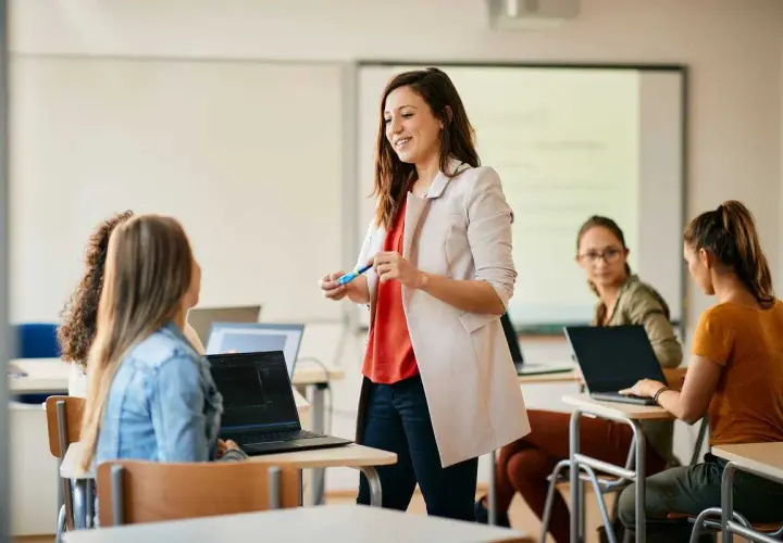Uśmiechnięta kobieta, w trakcie przygotowania do zawodu nauczyciela, prowadzi lekcję dla grupy studentek przy laptopach.