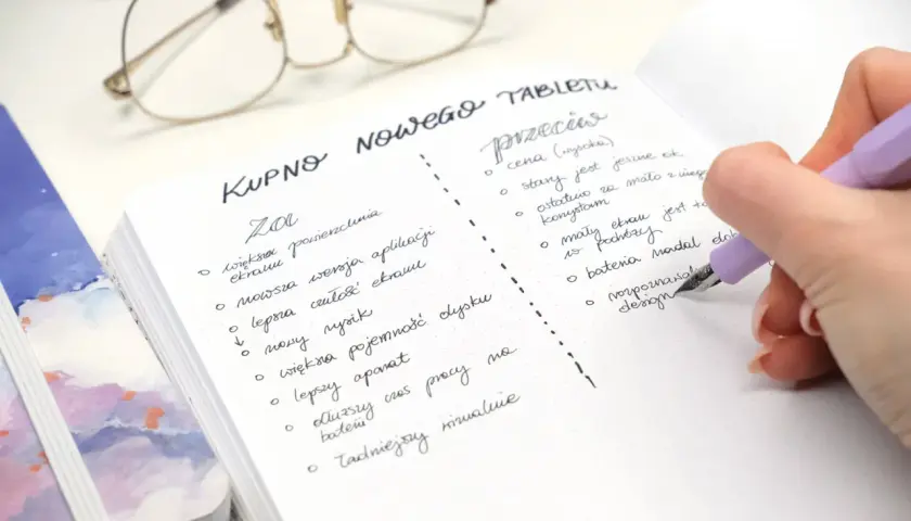 Ręka pisze notatkę w zeszycie: "Kupno nowego tabletu". Widoczne okulary i fragment innego notatnika.