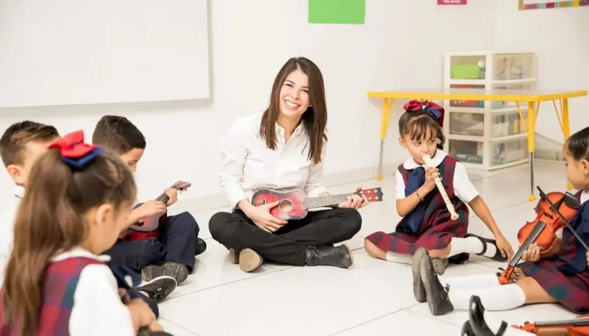 Uśmiechnięta nauczycielka gra na ukulele z dziećmi. To świetny przykład, jak zostać wychowawcą kolonijnym, ucząc przez zabawę.