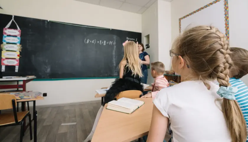 Dzieci w klasie, nauczycielka przy tablicy, gdzie widnieje zadanie matematyczne. Widać też tablicę z zadaniami, która może być częścią systemu motywacyjnego, np. dodatek za wychowawstwo.