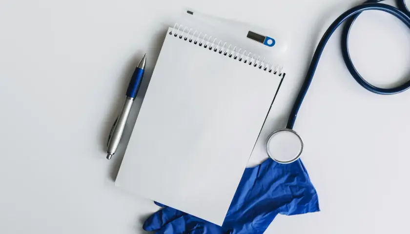 Lekarz przygotowuje dokumentację medyczną, notując informacje dotyczące usprawiedliwiania nieobecności w szkole rozporządzenie. Na stole leżą stetoskop, termometr, notatnik i rękawiczki.