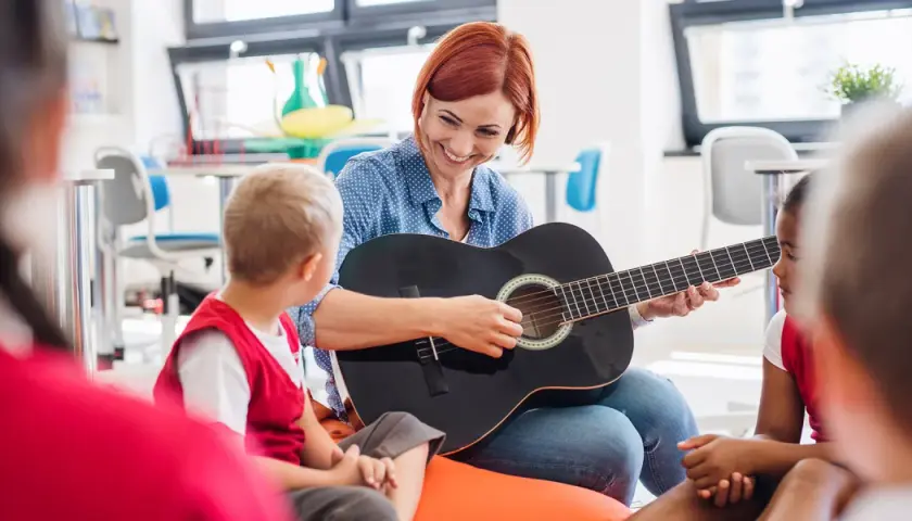 Nauczycielka gra na gitarze dla dzieci, tworząc zindywidualizowaną ścieżkę kształcenia. Przykłady takiej nauki to muzyka.