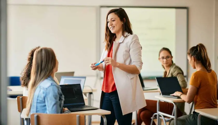 Uśmiechnięta kobieta, w trakcie przygotowania do zawodu nauczyciela, prowadzi lekcję dla grupy studentek przy laptopach.