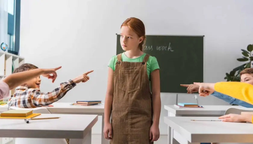 Smutna dziewczynka w brązowej sukience stoi sama, podczas gdy inne dzieci wskazują na nią palcami. To obrazek, który mógłby posłużyć jako opinia o uczniu do sądu, pokazując jego izolację.