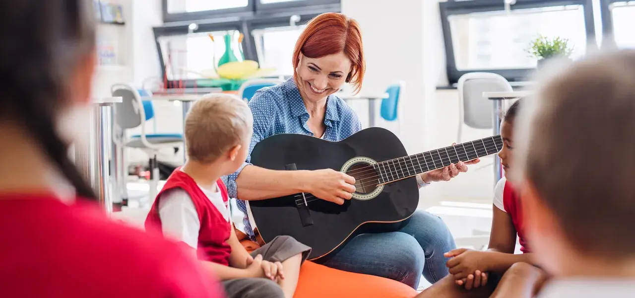 Nauczycielka gra na gitarze dla dzieci, tworząc zindywidualizowaną ścieżkę kształcenia. Przykłady takiej nauki to muzyka.