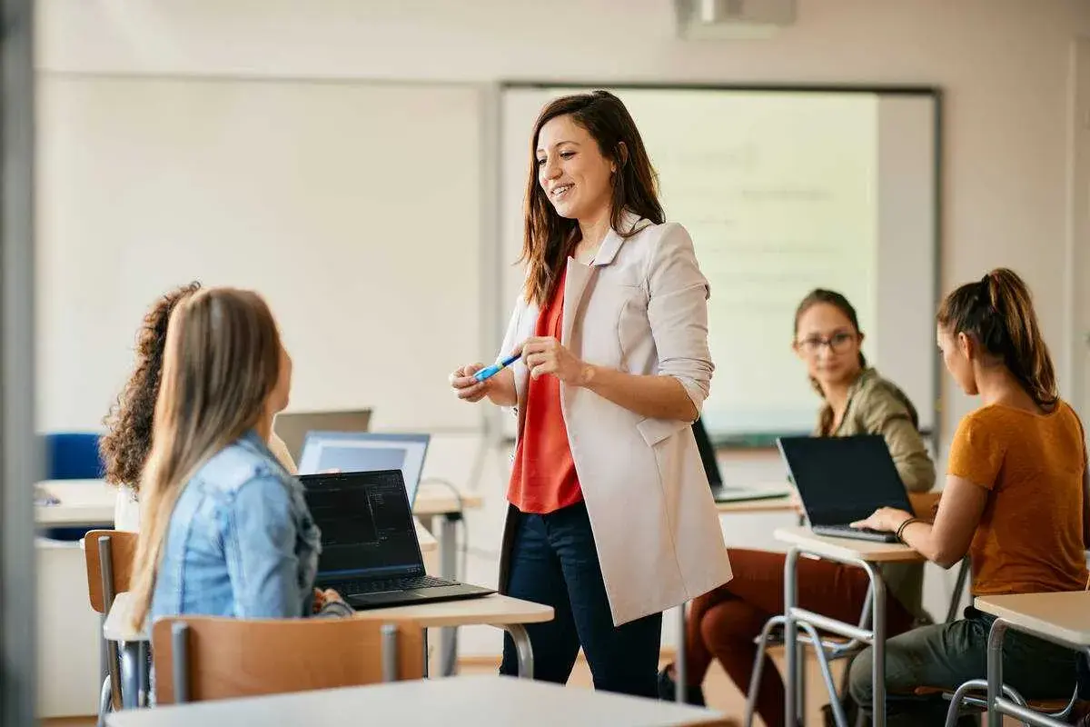 Uśmiechnięta kobieta, w trakcie przygotowania do zawodu nauczyciela, prowadzi lekcję dla grupy studentek przy laptopach.