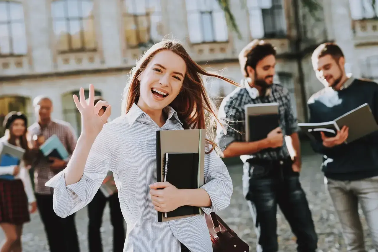 Uśmiechnięta studentka z notatnikami pokazuje znak OK. Pokazuje, jakie są perspektywy dla osób z wykształceniem humanistycznym – świetlane!