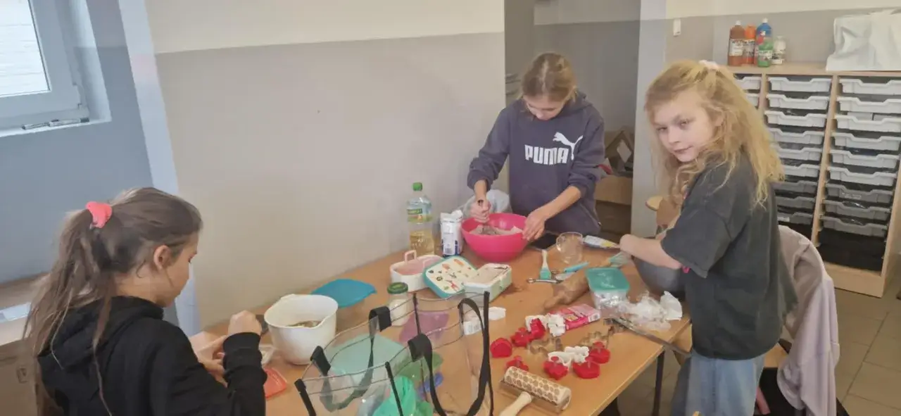 Dzieci w nowoczesnej klasie szkolnej przygotowują ciasto. Na stole stoją miski, składniki i foremki do ciastek.