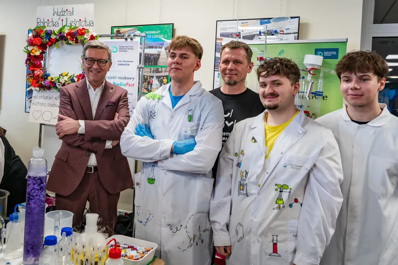Uśmiechnięty mężczyzna w garniturze i młodzi ludzie w kitlach laboratoryjnych pokazują, co można robić po dziennikarstwie – naukowe eksperymenty i innowacje.