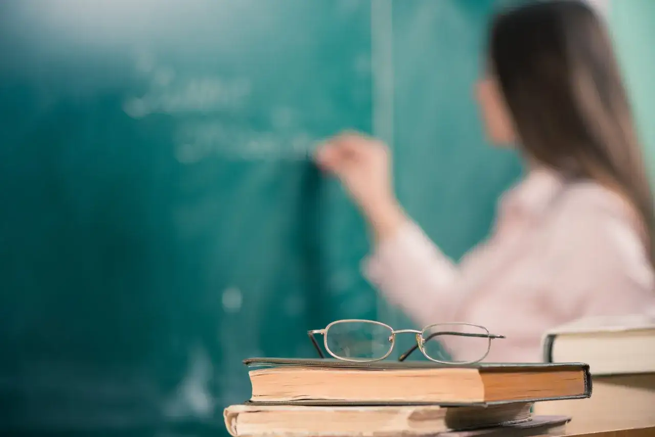 Okulary na książkach przed zieloną tablicą, gdzie nauczycielka pisze. To inspiracja dla klasy integracyjnej przepisy.