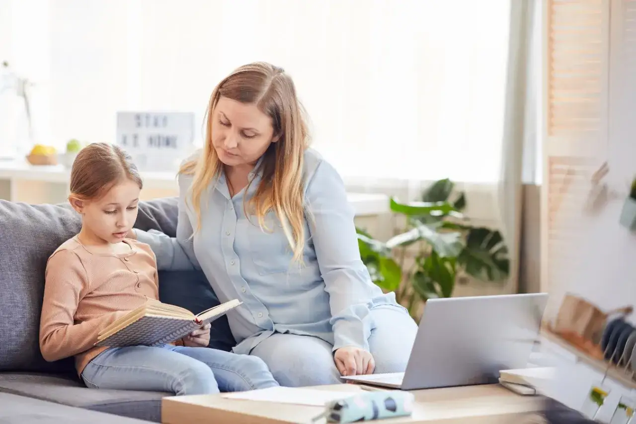 Mama wspiera córkę w nauce w domu, gdy ta ma nieobecność w szkole. Na stole leży laptop.