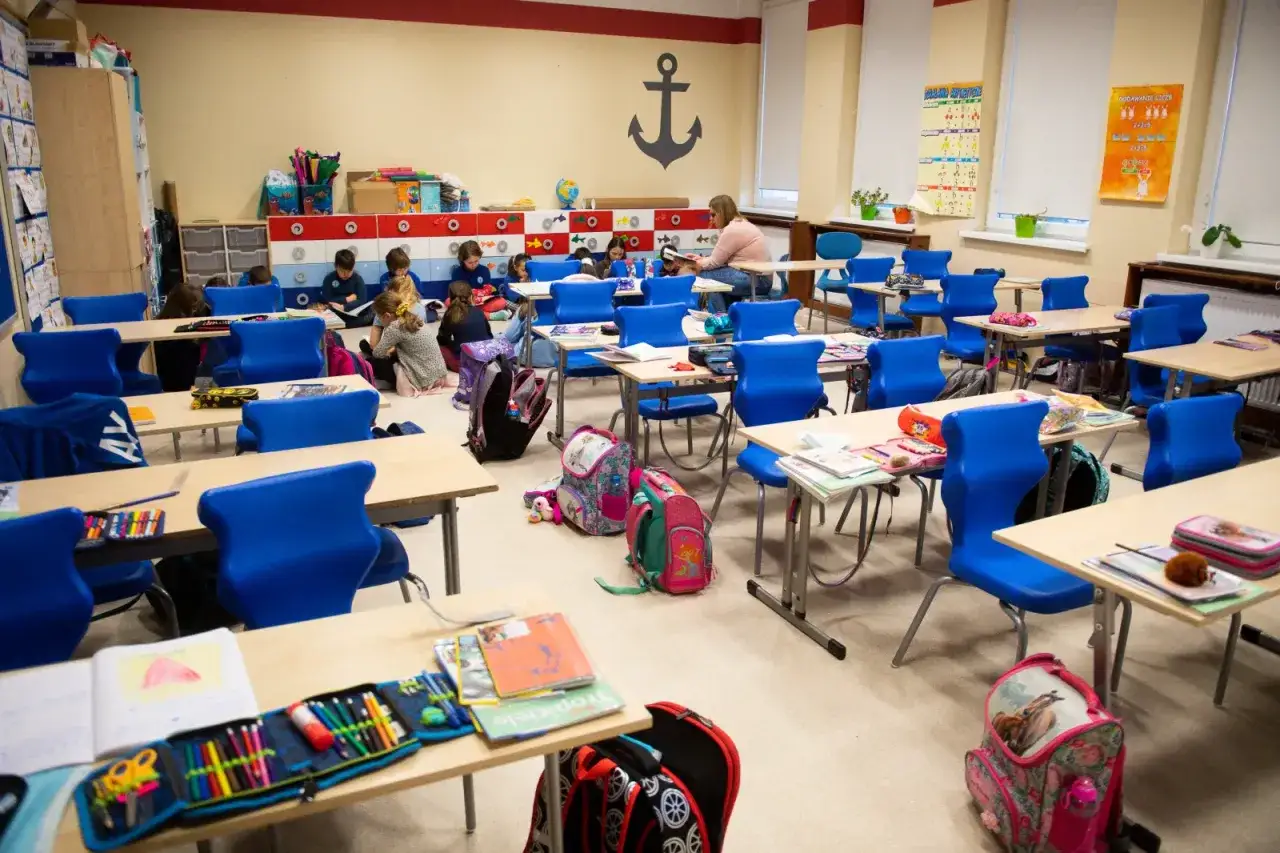 Klasa z dziećmi siedzącymi na podłodze i przy ławkach. Nauczycielka czyta książkę. Widoczna minimalna frekwencja w szkole, wiele pustych krzeseł.