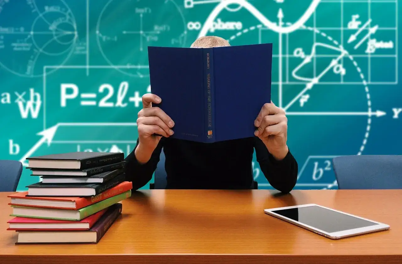 Osoba czyta książkę, przygotowanie do zawodu nauczyciela. Na stole stos książek i tablet. Tło z wzorami matematycznymi.