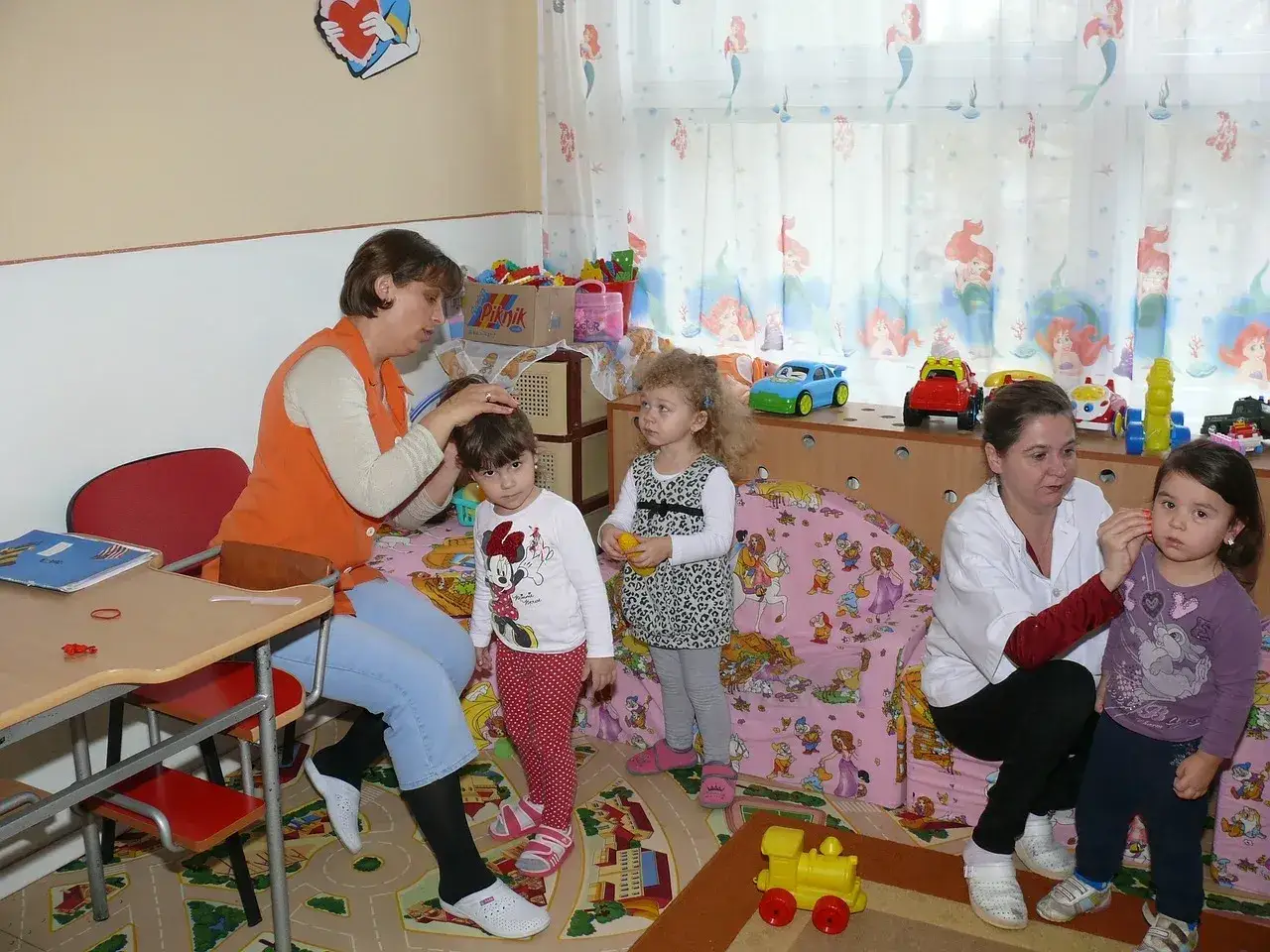 W przedszkolu od ilu lat? Nauczycielka czesze dziewczynkę, obok stoi koleżanka. W tle zabawki i bajkowe zasłony.