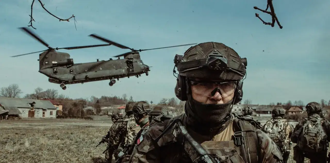 Żołnierze w pełnym rynsztunku, z helikopterem w tle. Chcesz wiedzieć, jak awansować w wojsku? Ciężka praca i determinacja to klucz.
