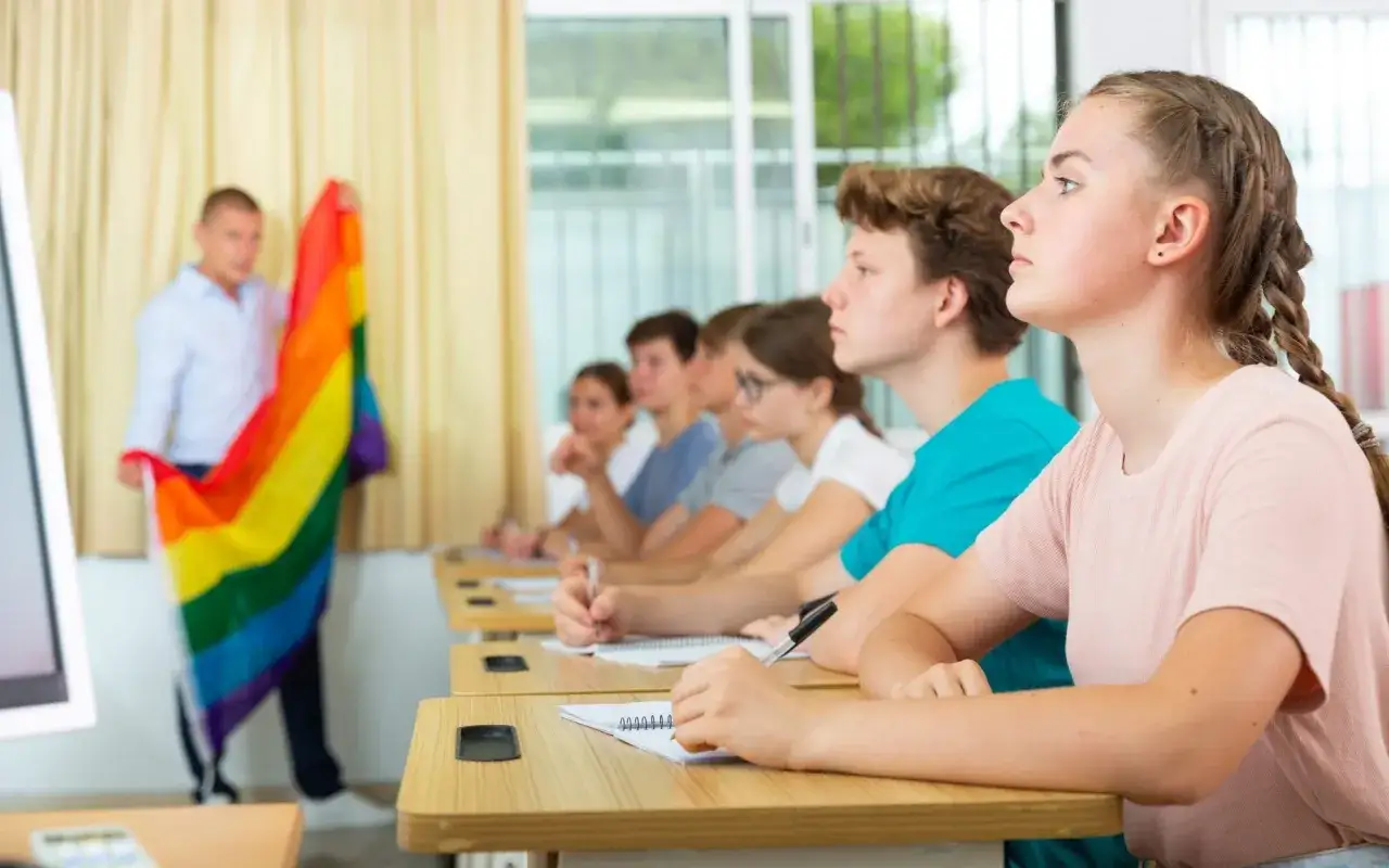 Nauczyciel prezentuje tęczową flagę, symbol akceptacji, podczas lekcji edukacji seksualnej w szkole. Uczniowie uważnie słuchają.