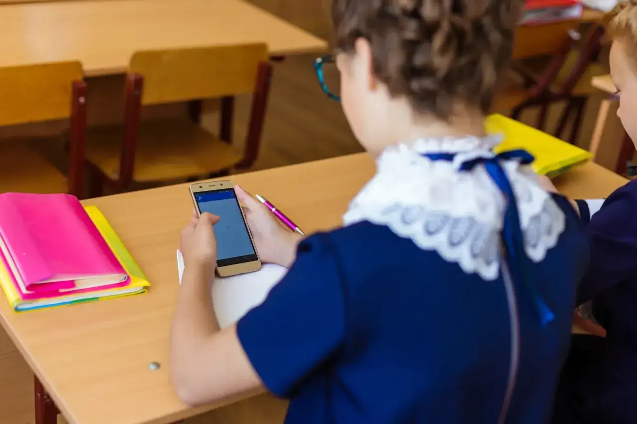 Uczennica w szkolnym mundurku trzyma telefon, zastanawiając się, czy nauczyciel może zabrać jej urządzenie. Obok leżą kolorowe książki.