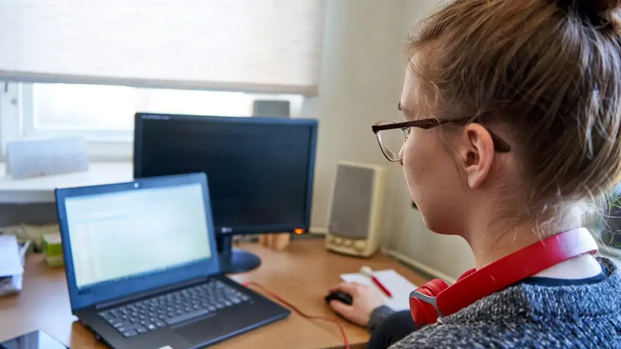 Dziewczyna w okularach pracuje przy laptopie, słuchawki na szyi. To obrazek z czasów nauki hybrydowej, gdy rozporządzenie pozwalało na elastyczność.