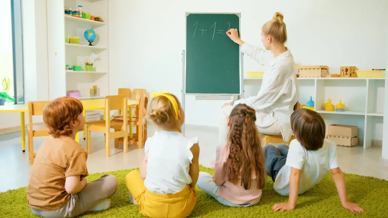 Nauczycielka pisze na tablicy, dzieci słuchają. Czy po kursie można pracować w przedszkolu? Tak, to świetny początek!